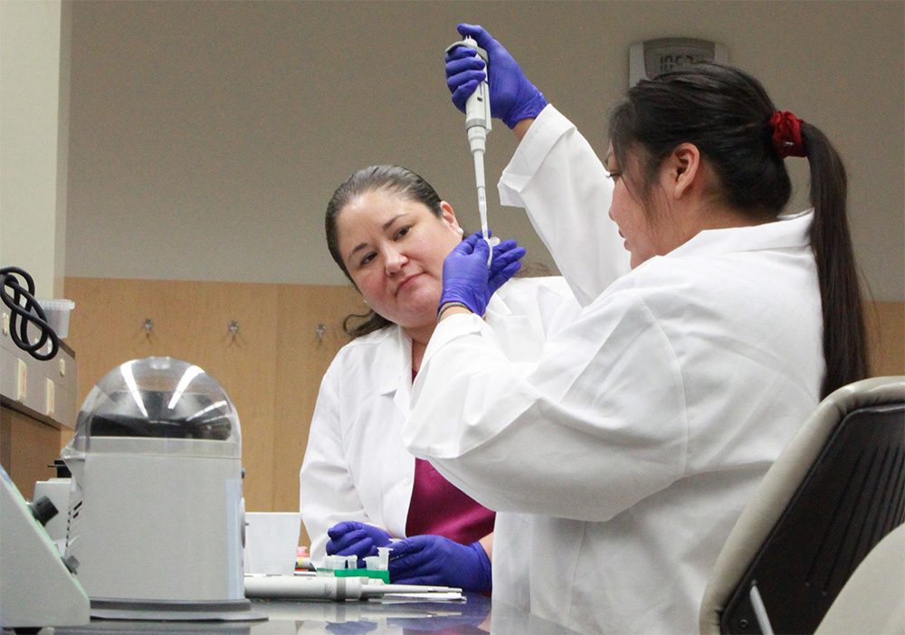 Johns Hopkins post-doctoral researcher Dr. Jessica L. Elm, citizen of the Oneida Nation and descendant of the Stockbridge-Munsee Band of the Mohicans, watches Alison Watson of the Navajo Nation as she receives hands-on training in genomics research at the 2019 SING workshop taking place on the University of Illinois Urbana campus.